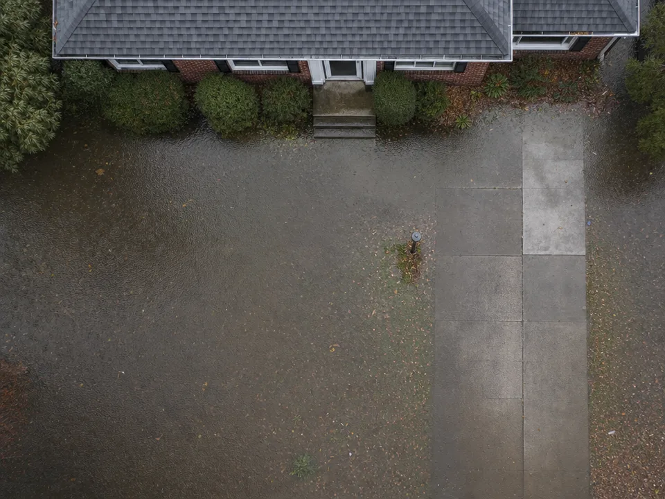 High overhead view of a brick ranch house's front yard and driveway under shallow brown floodwater, rain dimpling the surface