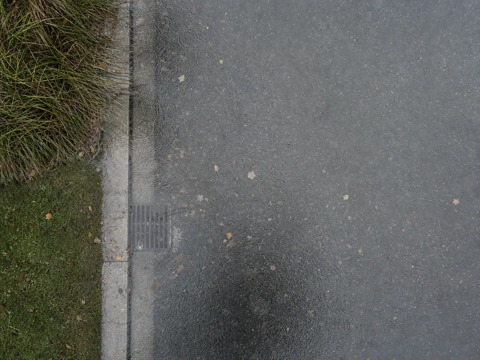 High overhead view of a street curb and storm drain under a thin sheet of standing water dimpled by rain, with grass and lawn along one edge