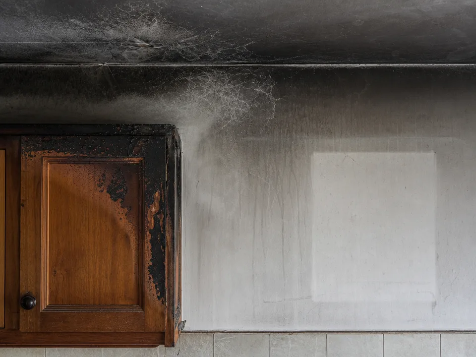 A kitchen wall and ceiling seen straight on, streaked with black soot above a scorched cabinet, a clean rectangle where a picture frame once hung